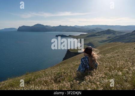 Frau hat Landschaft, Frau sitzt auf grasbewachsenem Hügel mit Blick auf Bucht und Berge an einem sonnigen Tag und genießt das friedliche Reiseziel Stockfoto