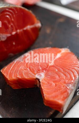 Makroansicht von zwei rohen geräucherten Sockeye-Lachssteaks, die auf einem dunklen Holzschneidebrett platziert sind, zeigt reiches orange-rotes Fleisch, sichtbare Muskelstränge, eine feuchte Oberfläche und einen dünnen silbernen Hautrand. weiches Naturlicht Stockfoto