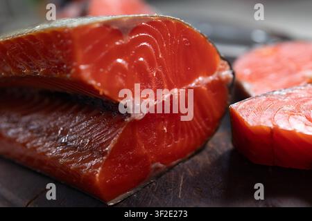 Makroaufnahme eines geräucherten Sockeye-Lachsfilets, das glänzendes tiefrotes Fleisch, feine Muskelmuster und dick geschnittene Portionen zeigt, die auf einem dunklen rustikalen Holzschneidebrett ruhen, mit weicher Hintergrundunschärfe Stockfoto