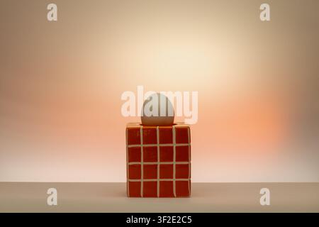 Konzeptionelle Stillleben mit rotem Gitterwürfel und grauem Ei auf einem warmen orange-pfirsichfarbenen Hintergrund. Weiche Studiolichtung, minimalistische herbstfarbene Komposition Stockfoto