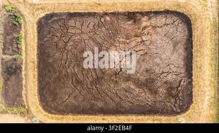 Luftaufnahme eines ausgetrockneten rechteckigen Teiches mit tiefen Rissen im verdorrten Erdreich, umgeben von trockenem Gras in Serbien. Stockfoto