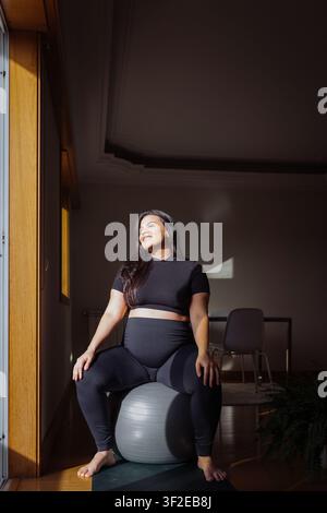 Lächelnde schwangere Frau in schwarzer Sportkleidung sitzt auf einem Fitnessball in einem sonnendurchfluteten Heimstudio und übt Pränatalyoga und achtsames Atmen für das Wohlbefinden Stockfoto