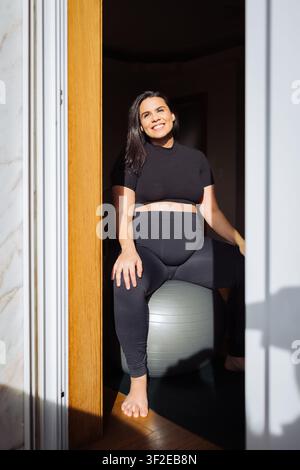 Lächelnde schwangere Frau in schwarzer Activewear sitzt auf einem Fitnessball zu Hause, beleuchtet von warmem Sonnenlicht durch eine Türöffnung. Fitness und pränatales Wohlbefinden co Stockfoto