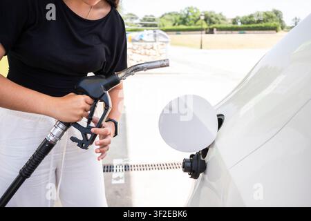 Nahaufnahme einer Frau, die einen Kraftstoffschlauch hält, während sie ein weißes Auto mit Diesel an einer Tankstelle betankt, wobei der Pumpengriff, der Schlauch und der offene Kraftstoffdeckel zu sehen sind Stockfoto