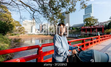 Eine ruhige NachmittagsSzene im Tennoji-Park von Osaka, mit einer Frau mit einem Kinderwagen auf der ikonischen roten Brücke, umgeben von urbaner Skyline und Natur Stockfoto