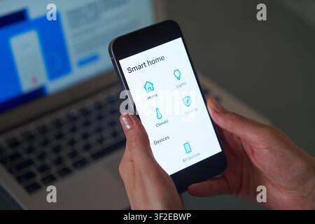 Eine Nahaufnahme von Händen, die ein Smartphone halten, das eine Smart-Home-Anwendungsschnittstelle anzeigt, mit Bedienelementen für Haus, Beleuchtung, Klima, Sicherheit und d Stockfoto