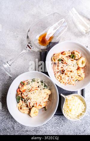 Oberblick auf zwei Schüsseln Tortellini-Pasta in Tomatensauce, mit geriebenem Käse und Basilikum garniert, serviert mit einem Glas Rotwein auf einem grauen Tisch, Italia Stockfoto