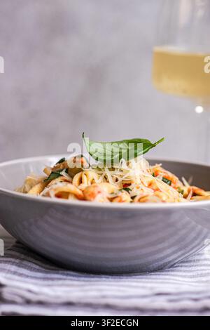 Nahaufnahme von cremigen Tortellini-Nudeln, überzogen mit geriebenem Parmesan und einem frischen Basilikumblatt in einer weißen Schüssel, serviert auf einem Tischgedeck mit weicher Unschärfe. Stockfoto