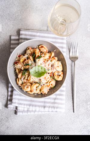 Oberblick auf Tortellini-Pasta, die mit Tomatensauce, frischem Basilikum und geriebenem Parmesan in einer Keramikschüssel auf einem gestreiften Geschirrtuch geworfen wurde, mit Gabel und wh Stockfoto