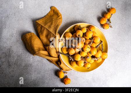 Oberansicht von reifen Loquats in einer gelben Schale mit rustikalem Serviette auf grauer Steinoberfläche, gesundes frisches Produktkonzept reich an Vitamin C. Stockfoto