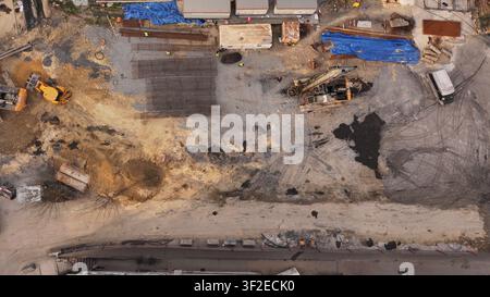 Luftbild von oben einer aktiven Baustelle mit Bagger, Lader, Lastwagen und gestapelten Bewehrungsmatten auf Kiesboden, das industrielle Arbeit zeigt Stockfoto