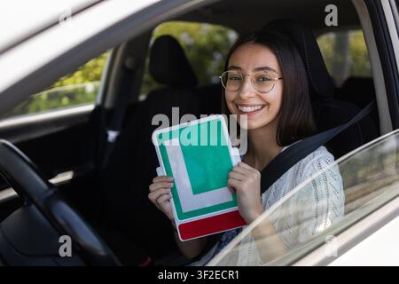 Lächelnde junge Frau mit Brille sitzt auf dem Fahrersitz mit angezogener Sicherheitsgurt, hält ein L-Plakettenschild durch ein offenes Autofenster an einem sonnigen Tag. Stockfoto