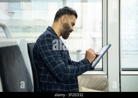 Seitenansicht eines indischen Mannes, der mit einer Stadtbahn pendelt und am hellen Fenster sitzend mit einem Tablet und einem Stift liest, schreibt oder Arbeit plant. Stockfoto