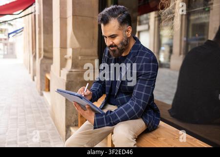 Indischer Geschäftsmann in einem karierten Blazer sitzt auf einer Holzbank vor einem städtischen Gebäude, schreibt auf einem Tablet mit einem Stift und konzentriert sich auf die Arbeit und Plansi Stockfoto