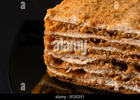 Nahaufnahme einer geschichteten Honigkuchenscheibe mit cremiger Füllung und Krümel-Topping auf einem dunklen Teller. Reichhaltige Bäckerei-Desserttextur im warmen Licht gezeigt, perfekt Stockfoto