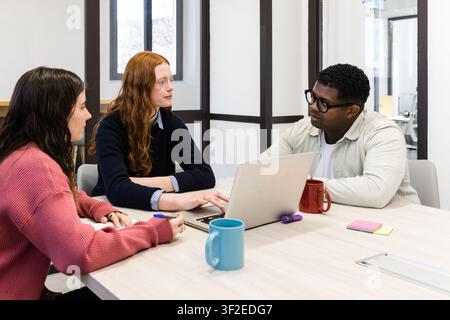 Verschiedene Kollegen arbeiten gemeinsam an einem Besprechungstisch in einem modernen Büro, diskutieren Ideen über einen Laptop, Notizbücher und Kaffeetassen und betonen die Teamarbeit Stockfoto