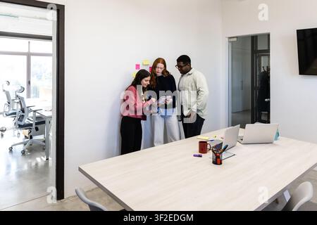Drei Kollegen arbeiten in einem modernen Bürobesprechungsraum zusammen, überprüfen Notizen auf einem Tablet in der Nähe einer weißen Wand mit Haftnotizen, planen Aufgaben und Ideen Stockfoto