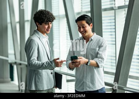 Zwei Geschäftskollegen lächeln, während sie Informationen auf einem Smartphone in einem hellen modernen Bürokorridor überprüfen und über ein Projektupdate und die Planung diskutieren Stockfoto