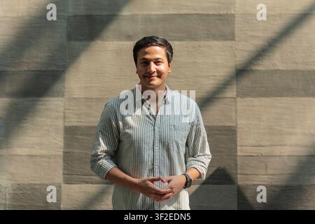 Porträt eines lächelnden Mannes in einem gestreiften Hemd, der vor einer Steinmauer steht, beleuchtet von natürlichen Fensterschatten. Entspannte Haltung mit verschränkten Händen, urban indo Stockfoto