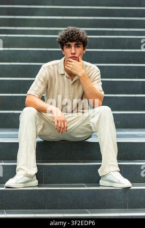 Porträt eines jungen Mannes, der auf städtischen Treppen sitzt, das Kinn auf die Hand gestützt in einer nachdenklichen Pose, der lässige helle Kleidung in einer modernen Stadt trägt Stockfoto