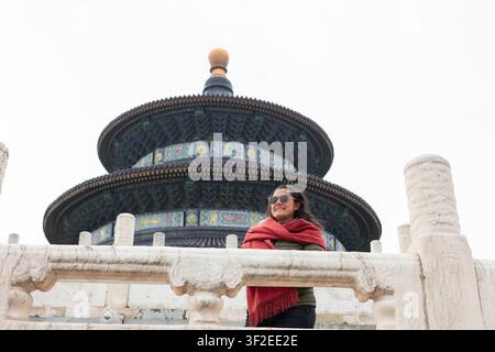 Reisender mit Sonnenbrille und rotem Schal lächelt am Tempel des Himmels in Peking, China, an einem bewölkten Tag und hebt historische Architektur und Kult hervor Stockfoto