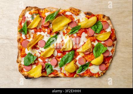 Italienische Gourmet-Pinsa Romana mit geschnittenem Wurstel, gebratenen Kartoffeln, Tomatensauce, geschmolzenem Mozzarella und frischem Basilikum, serviert auf einem Holzbrett Stockfoto