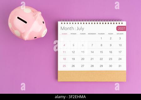 Juli 2027 Tischkalender und rosa Sparschwein auf violem Farbhintergrund. Stockfoto