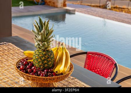 Ansicht eines Korbs gefüllt mit einer frischen Ananas, reifen Bananen und dunkelroten Kirschen auf einem Tisch neben einem Schwimmbad in Chania, Griechenland. Stockfoto