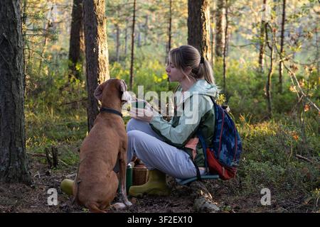 Positive Frau Backpackerin wandert mit Hund im Wald, sitzt auf einem Baumstamm, entspannt sich und isst Snacks. Stockfoto