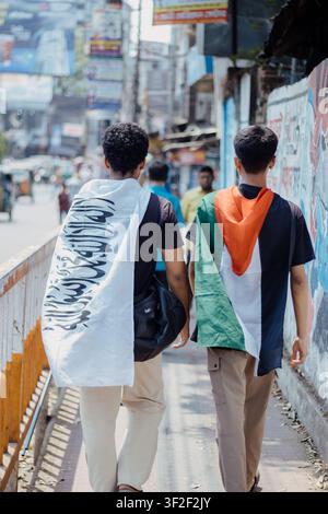 Feni, Bangladesch - 07. April 2025: Blick auf zwei junge Männer, die eine Straße in Feni entlanggehen, einer trägt eine Flagge mit arabischer Schrift und der andere trägt eine Stockfoto