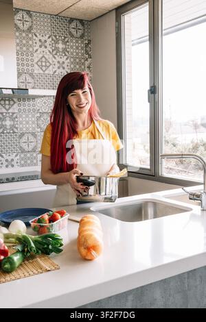 Frau lächelt, während sie einen Topf mit Nudeln hält, in einer modernen Küche stehend Stockfoto
