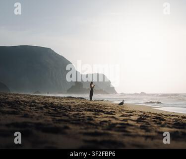 Distrikt Faro, Portugal - 20. September 2024: Blick auf eine Frau, die auf einem Sandstrand in der Nähe des Ozeans steht, mit dramatischen Klippen im Hintergrund während go Stockfoto