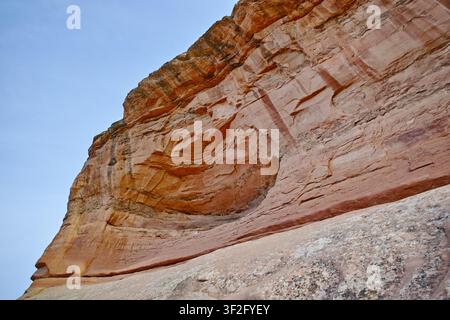 Schichtiger roter Sandsteinklippe erhebt sich vor einem hellblauen Himmel mit verwitterten Texturen. Stockfoto