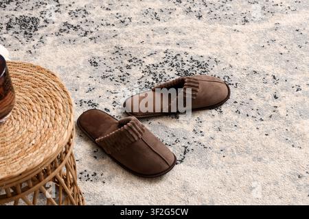 Paar braune Wildlederpantoffeln auf einem strukturierten grau-weißen Teppich neben einem geflochtenen Rattan-Beistelltisch, Komfort und Entspannung in einem Wohninterieur se Stockfoto