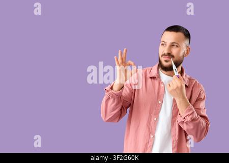 Hübscher junger Mann mit Thermometer, das OK-Geste auf Flieder-Hintergrund zeigt Stockfoto