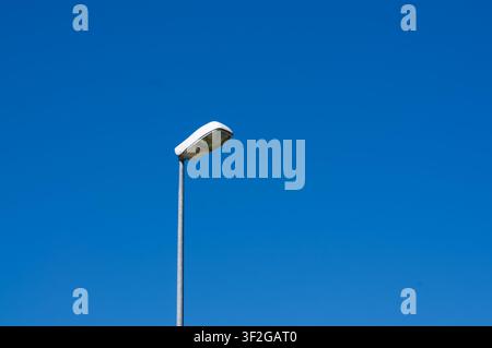 Niedrig angelegte Ansicht einer modernen öffentlichen Straßenlaterne, die auf einem geraden Metallmast montiert ist und sich auf der linken Seite befindet. Der Laternenmast steht vor blauem Himmel Stockfoto