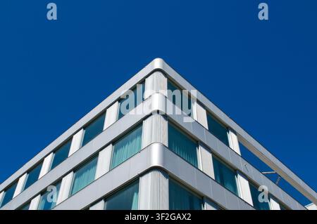 Niedrigwinklige Ansicht einer Ecke eines zeitgenössischen Bürogebäudes, das abstrakte geometrische Architektur mit silberner Metallverkleidung und großen Glasfenstern zeigt. Stockfoto
