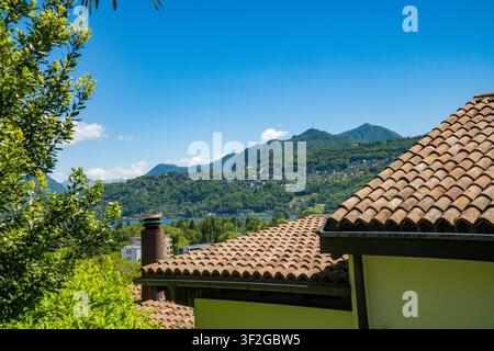 Traditionelle Dachplatten aus Terrakotta mit Schornstein, Blick auf den fernen See und die Berge, eingerahmt von grünen Bäumen, Tessin, Schweiz. Stockfoto