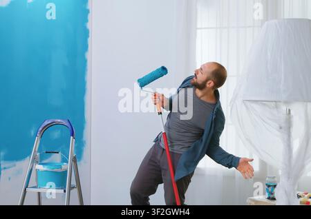 Zufriedener Handwerker singt in eine Walzenbürste während der Renovierung einer Wohnung, verwendet blaue Farbe und verwandelt die Wohnungseinrichtung in einen verspielten DIY-Moment. Stockfoto