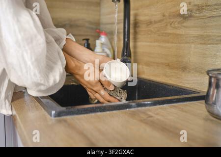 Person wäscht Tasse im Küchenwaschbecken. Wasser läuft aus dem Wasserhahn, während Hände Tasse und Schwamm halten. Einfache Aufgabe, die in der Küchenarbeitsplatte mit Holzoberflächen stattfindet und Stockfoto