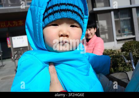 Ein Baby in einem blauen Kapuzenmantel und einer Strickmütze wird im Freien gehalten, während eine Frau im Hintergrund steht. Stockfoto