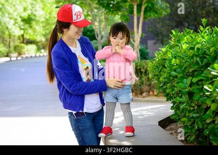 Eine Frau in einer blauen Jacke hält ein Kind in Rosa neben grünen Sträuchern auf einem gepflasterten Weg. Stockfoto