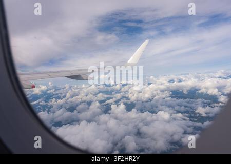 Flugzeugflügel sichtbar durch ein Fenster, während man über weißen, flauschigen Wolken fliegt, mit einem malerischen Blick auf den Himmel und eine weite, wolkige Landschaft darunter Stockfoto