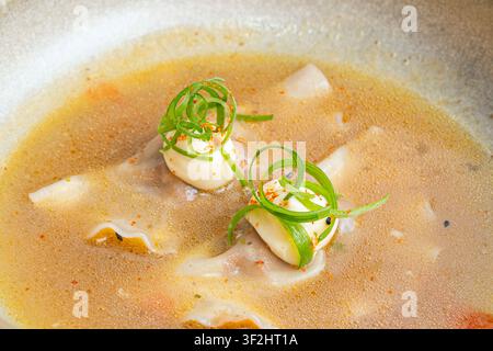 Nahaufnahme von Knödeln, die in sprudelnder bernsteinfarbener Brühe schwimmen, belegt mit Sahnetropfen, spiralförmigen Frühlingszwiebel-Curls, Chiliflocken und schwarzen Sesamsamen. Stockfoto