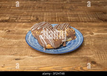 Trio von schokoladenüberzogenen Herzgebäckstücken mit weißem Zuckerguss und zerkleinerten Nüssen auf einem blauen Keramikteller über einem Holztisch. Stockfoto