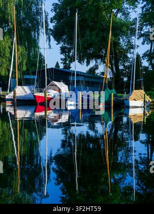 Farbenfrohe Segelboote, die während der goldenen Stunde auf ruhigem Wasser vor Anker liegen, mit lebhaften Spiegelungen und üppigen grünen Bäumen im Hintergrund. Reflektierte Boote, Spiegel. Stockfoto