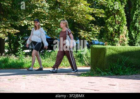 Kiew, Ukraine – 17. Juli 2026: Zwei Frauen gehen einen schattigen Parkweg entlang, umgeben von grünen Bäumen am VDNH (Expocenter) an einem sonnigen Sommertag. Stockfoto