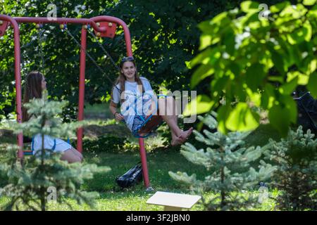 Kiew, Ukraine - 17. Juli 2026: Eine junge Frau entspannt sich auf einer Spielplatzschaukel, während sie Zeit mit einer Freundin in einem schattigen Parkbereich am VDNH (Expocenter) verbringt Stockfoto