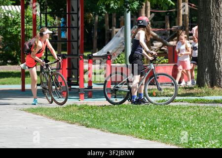 Kiew, Ukraine - 17. Juli 2026: Besucher laufen mit Fahrrädern und verbringen Zeit in der Nähe eines Spielplatzes und eines Freiluft-Erholungsgebiets am VDNH (Expocenter) an einem sonn Stockfoto