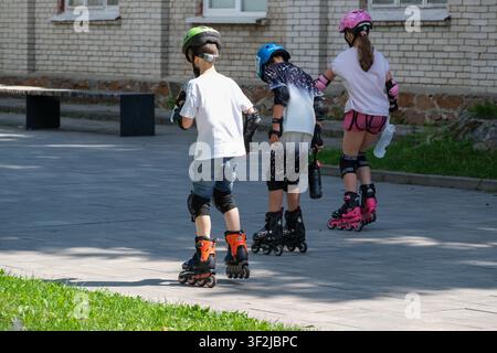 Kiew, Ukraine - 17. Juli 2026: Drei Kinder, die Helme und Schutzausrüstung tragen, üben Inlineskaten gemeinsam in einem Freizeitareal bei VD Stockfoto